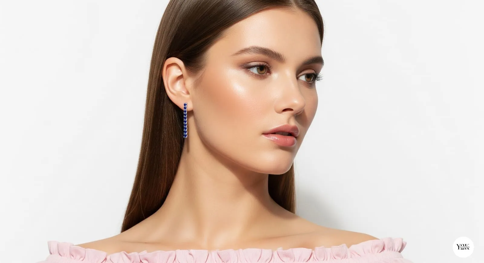 A woman in a soft pink off-the-shoulder top, wearing long, dangling Sapphire Tennis earrings. Her neutral makeup and poised expression highlight the simplicity and elegance of the jewelry.