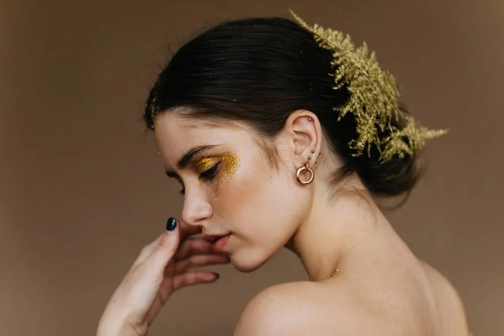 A woman adorned with delicate gold hoop earrings and a matching gold headpiece, featuring intricate floral details. Her golden eye makeup and dark lipstick add to the glamorous and festive look.